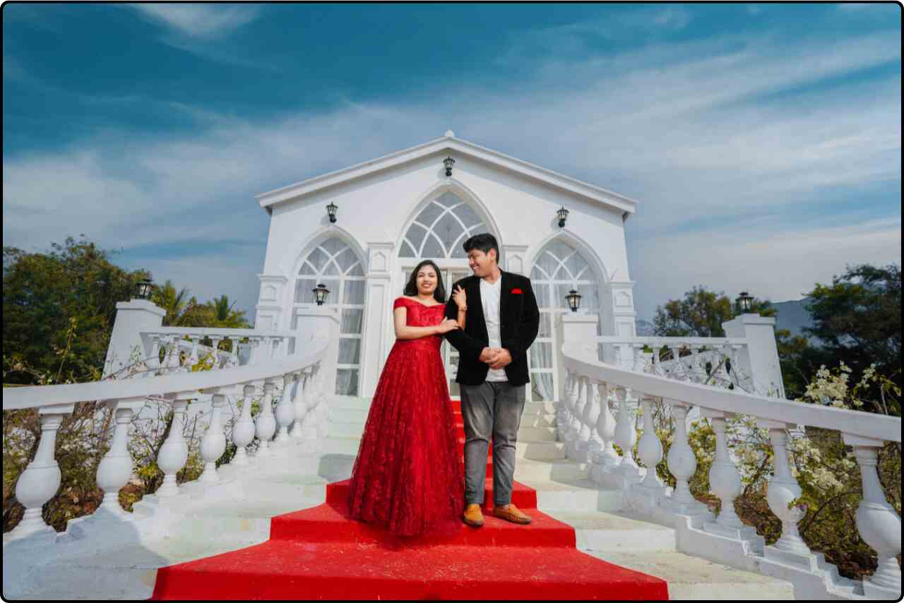 Newlyweds standing on ornate stairs, the bride in a red dress and the groom in a black tuxedo, background view is  Charge of Jesus Christ.